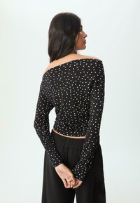 Schwarzes Langarmoberteil mit weißen Punkten. Off-Shoulder-Design, figurbetonte Form und weicher Stoff; kombiniert mit lockeren schwarzen Hosen.