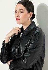 Musta nahkatakki sileällä pinnalla, jossa on vetoketjulukko ja klassinen kaulus. Malli poseeraa käsi leuan lähellä, esitellen muotoilua.