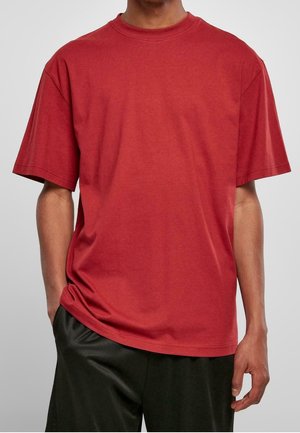 Mężczyzna w prostym czerwonym T-shircie z krótkim rękawem i czarnych spodniach, z jedną ręką w kieszeni, na jednolitym jasnoszarym tle.