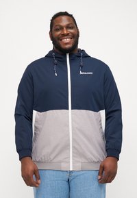 Chaqueta ligera en bloque de colores azul marino y gris, con capucha, cierre de cremallera en la parte delantera, puños elásticos y detalles con cordón. Logo de Jack & Jones en el pecho.