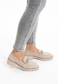 Beige suede loafers with a chain accent, featuring a rugged outsole and contrasting stitching, paired with slim grey jeans.