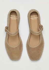 Chaussures en daim beige avec une pointe arrondie, une sangle avec une boucle et un détail brodé le long de la semelle. Les étiquettes indiquent "Femme par Fred Martin, Fabriqué en Espagne."