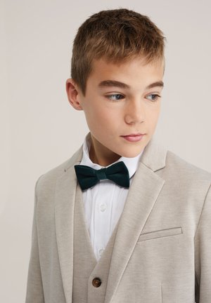 Blazer beige con finitura strutturata, gilet coordinato, camicia bianca e papillon in velluto verde. Presenta una piccola tasca sul petto e dettagli con bottoni.