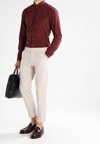 Muž oblečený v maroon košili na knoflíky, béžových kalhotách, tmavě hnědých mokasínech a držící černou texturovanou koženou aktovku.