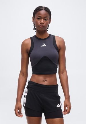 Joven mujer con top corto sin mangas Adidas en negro y gris y pantalones cortos negros Adidas con capas, de pie frente a un fondo liso y claro.