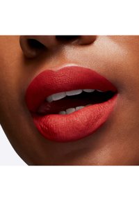 Rossetto opaco rosso applicato su labbra piene, che mette in mostra una texture liscia e contorni definiti, con denti visibili. Il tono della pelle appare caldo.