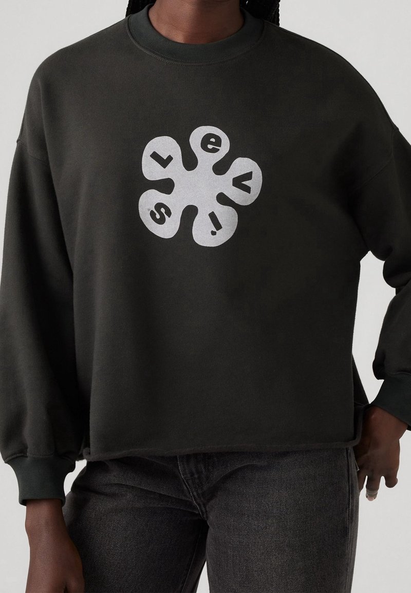 Sweat-shirt noir avec un motif floral abstrait blanc comportant les lettres "L", "e", "v", "i" et "s" sur la poitrine, porté avec un jean foncé.