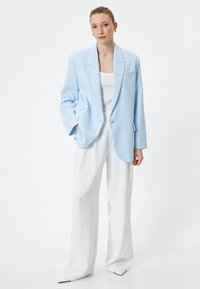 Blazer oversized blu chiaro con grandi rever, chiusura a singolo bottone e tasche laterali, abbinato a pantaloni bianchi a gamba larga e scarpe a punta.