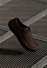 Scarpa slip-on in suede marrone con punta arrotondata, caratterizzata da un design minimalista, suola in gomma nera a contrasto e sottili dettagli di cucitura.