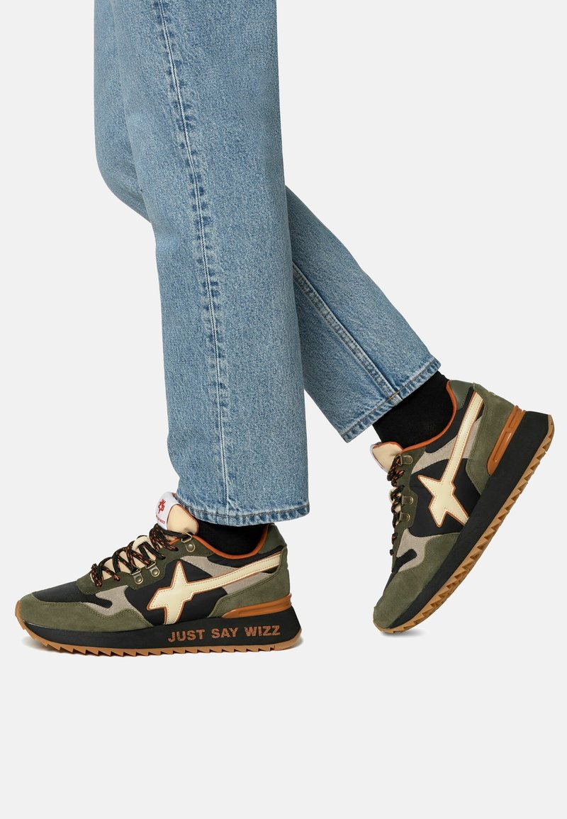 Personne portant un jean bleu clair et des baskets vertes, noires et beiges avec le texte "JUST SAY WIZZ" sur la semelle, marchant sur un fond blanc.