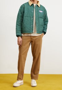 Grön kviltad jacka med beige krage, med en patchlogotyp. Bruna avslappnade byxor och krämfärgade sneakers med en strukturerad sula. Gul bakgrund.