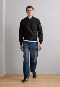 Schwarze Bomberjacke mit einem Reißverschluss vorne, eng anliegenden Bündchen und einer glatten Textur, kombiniert mit blauen, lässig aufgetragenen Jeans und schwarzen Sportschuhen.