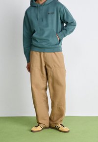 Sudadera con capucha de color verde azulado con bolsillo delantero y logo negro, combinada con pantalones de corte suelto color marrón claro que cuentan con bolsillos y detalles de costura, y zapatos amarillos.