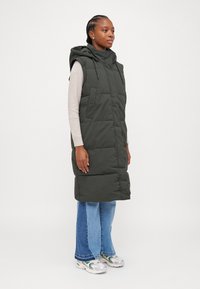 Polstret, ermeløs vest i mørkegrønn, med hette, sidelommer og knappelukking. Kombinert med en lys genser og utsvakt denim.