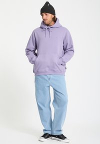 Hoodie violet avec une poche kangourou et des cordons, associé à un pantalon en denim bleu clair et des baskets noires. Le modèle porte un bonnet noir.