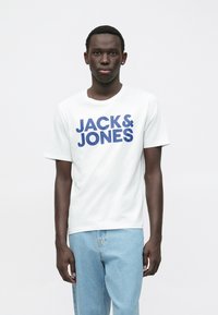 Ung mann iført en hvit T-skjorte med "JACK & JONES" i blå tekst og lyseblå jeans, stående mot en ensfarget lys bakgrunn.