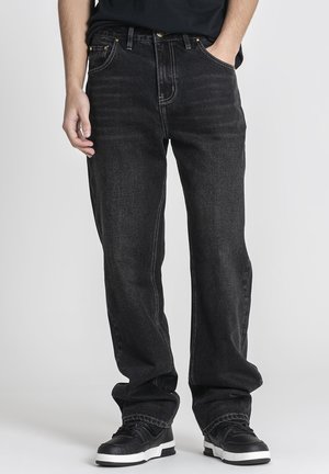 Mann trägt lockere schwarze Jeans und schwarze Sneakers mit weißen Sohlen, steht vor einem neutralen Hintergrund.