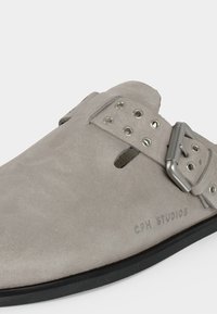 Ανοιχτό γκρι σουέντ slip-on παπούτσι με ρυθμιζόμενο μεταλλικό πόρπη, πολλαπλές τρύπες και μαύρη σόλα, με το λογότυπο "CPH STUDIOS" στο πλάι.