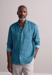 Camisa de botón color verde azulado con un patrón geométrico, que incluye un bolsillo en el pecho y mangas arremangadas. Combinada con pantalones de color claro.