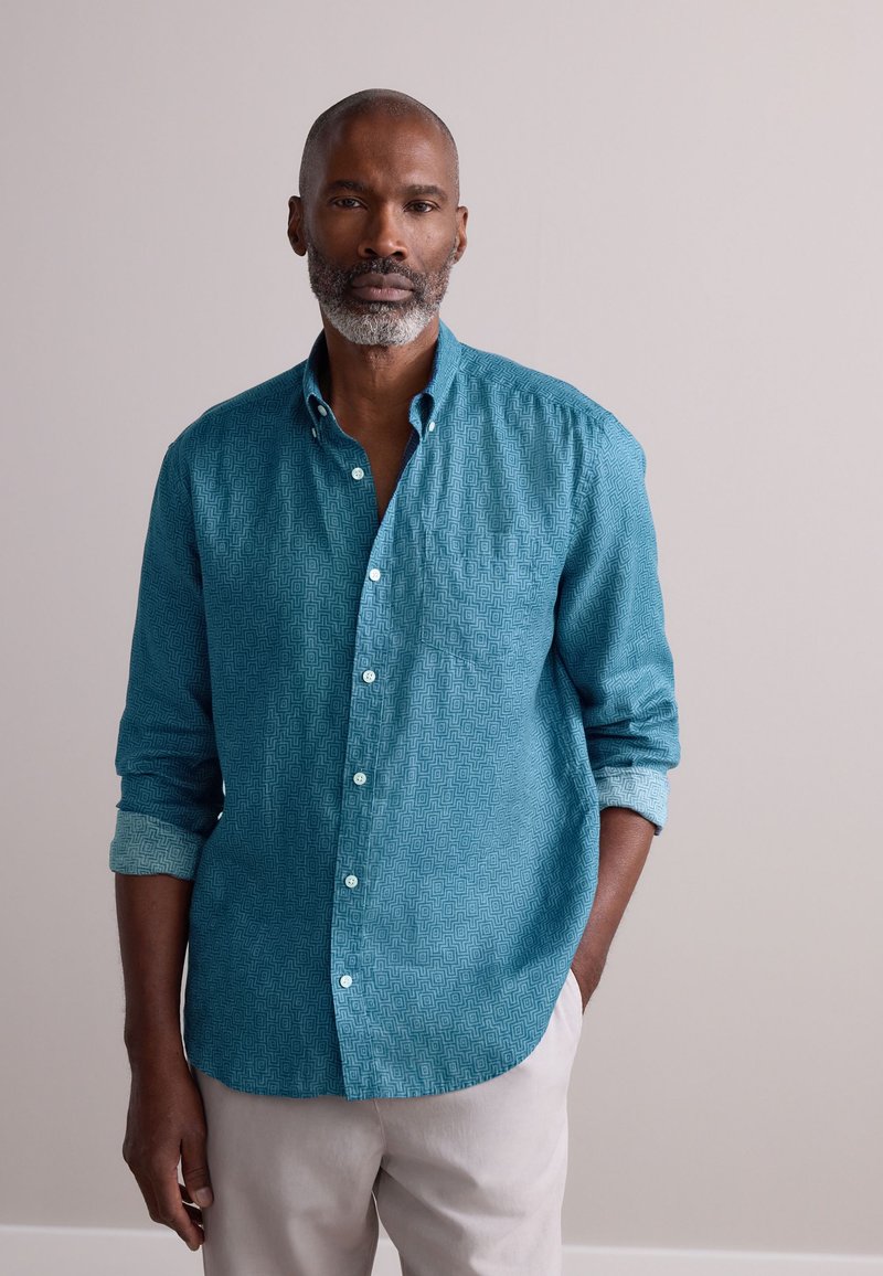 Camisa de botón color verde azulado con un patrón geométrico, que incluye un bolsillo en el pecho y mangas arremangadas. Combinada con pantalones de color claro.