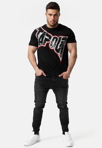 Camiseta de algodón negra con un gran logo desgastado de "TAPOUT" en blanco y rojo. Combinada con jeans negros ajustados y zapatillas blancas.