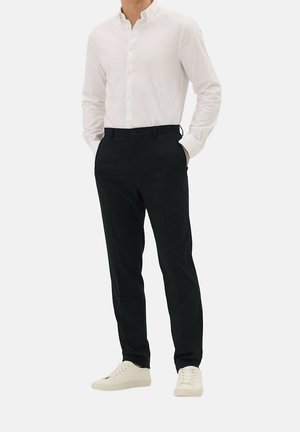 Mann trägt weißes Hemd mit Knöpfen, schwarze maßgeschneiderte Hose und weiße Turnschuhe, steht mit den Händen in den Taschen vor einem schlichten Hintergrund.
