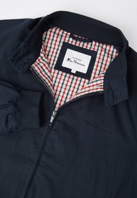 Navy bomber jacket with a ribbed collar, zip closure, and checkered red and white interior lining. Features embroidered logo on chest.