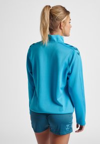 Lichtblauw sportshirt met lange mouwen en hoge kraag, met zwarte chevronaccenten op de schouders, in combinatie met donkerblauwe shorts. Soepele textuur.