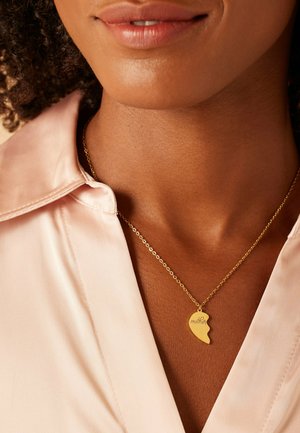 Collier en or avec un pendentif en forme de cœur gravé du mot "maman". La chaîne est délicate et complète le blouse rose pâle.