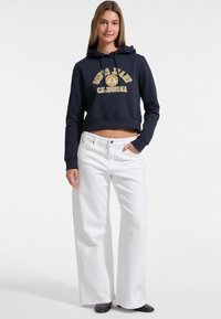 Felpa corta blu navy con scritte bianche, abbinata a jeans bianchi a gamba larga. Il tessuto sembra morbido e informale, con una vestibilità rilassata.