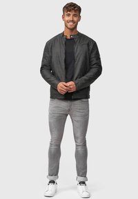Chaqueta bomber negra con una textura suave, cierre frontal y acentos de canalé, combinada con jeans gris slim-fit y zapatillas blancas.
