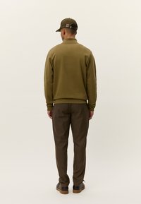 Sweatshirt long à manches en vert olive avec des poignets côtelés, associé à un pantalon à carreaux marron. Le mannequin porte une casquette assortie en olive.