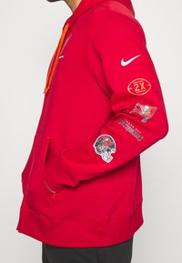 Rød Nike hoodie med orange snore, der har Tampa Bay Buccaneers logoer og grafik på ærmerne, forlommer og en blød stoftekstur.