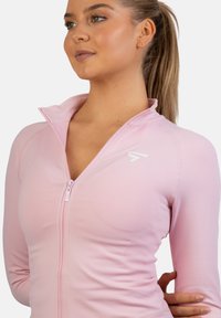 Lyserød atletisk zip-up jakke lavet af strækbart materiale, med høj krave og et lille hvidt logo på brystet.