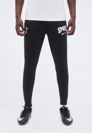 Partie inférieure du corps d'une personne portant un pantalon de sport noir des Spurs avec des logos, des chaussettes blanches et des crampons noirs avec des accents bleu clair, debout sur un fond blanc.