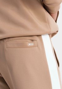 Gros plan sur un pantalon beige avec une bande latérale blanche et une poche passepoilée à l'arrière ornée d'une petite étiquette logo rectangulaire.