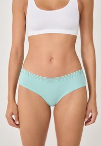 Frau in hellblauen Baumwoll-Hipster-Unterhosen und einem weißen Sport-BH, vor einem einfarbigen Hintergrund stehend.