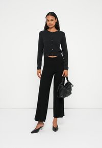 Μαύρος κοντός cardigan με κουμπί, σε συνδυασμό με φαρδιά μαύρα παντελόνια. Τα αξεσουάρ περιλαμβάνουν μια μαύρη υφασμάτινη τσάντα και κομψά τακούνια.