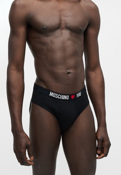 Άνδρας φοράει μαύρα εσώρουχα Moschino με λευκό κείμενο και κόκκινη καρδιά στη μέση, στέκεται μπροστά σε απλό φωτεινό φόντο.