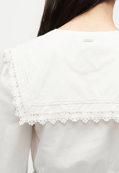 Blouse blanche avec large col carré présentant une bordure en dentelle complexe, portée par une personne aux longs cheveux foncés, vue de dos montrant une petite étiquette métallique de la marque.