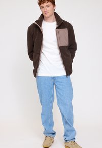 Brown fleece jacket with a tan zippered chest pocket, high collar, worn over a white t-shirt, paired with light blue jeans and beige shoes.