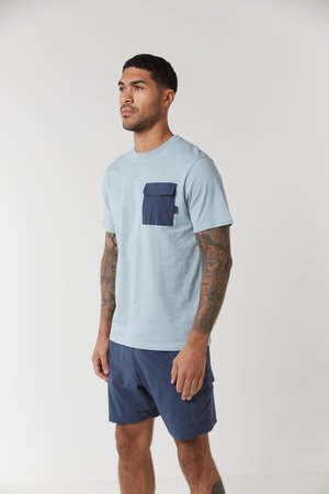 T-shirt azzurro chiaro con taschino blu navy, collo rotondo e maniche corte. Abbinato a pantaloni corti blu navy con tasche laterali.