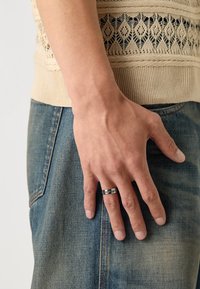 Sølvring med inngravert detaljer som bæres på pekefinger. Myk beige strikket topp og blå denimjeans er delvis synlige.