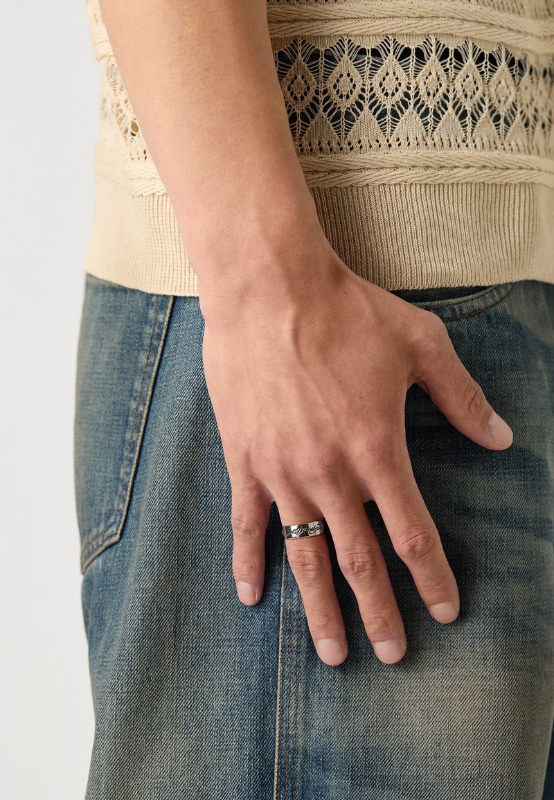 Sølvring med inngravert detaljer som bæres på pekefinger. Myk beige strikket topp og blå denimjeans er delvis synlige.