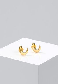 Pendientes de studs en forma de media luna dorada con un acabado pulido, que cuentan con un pequeño respaldo de poste. Colocados sobre una superficie de exhibición minimalista.