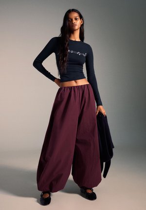 Čierne tričko s dlhým rukávom s grafickým dizajnom, skombinované s voľnými širokými nohavicami bordovej farby. Oblečené s čiernymi balerínkami a tmavým svetrom prehodeným cez rameno.