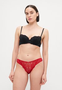 Zwarte kanten gepadde beha met verstelbare bandjes, gecombineerd met een rode kanten en mesh string met een kleine decoratieve accent aan de voorkant.