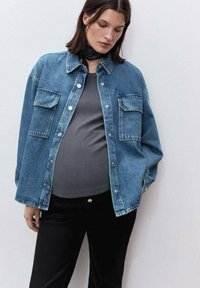 Giacca di denim azzurro chiaro con due tasche sul petto, bottoni silver e una vestibilità rilassata, indossata sopra una canotta grigia e pantaloni neri.