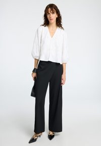 Blouse blanche avec des motifs texturés, décolleté en V et manches bouffantes, associée à un pantalon noir à jambes larges et des talons aiguilles noirs. Pochette noire.