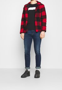 Rote und schwarze karierte Fleecejacke, schwarzes T-Shirt mit weißem Streifen, eng anliegende blaue Jeans und schwarze Sneakers mit weißen Socken.